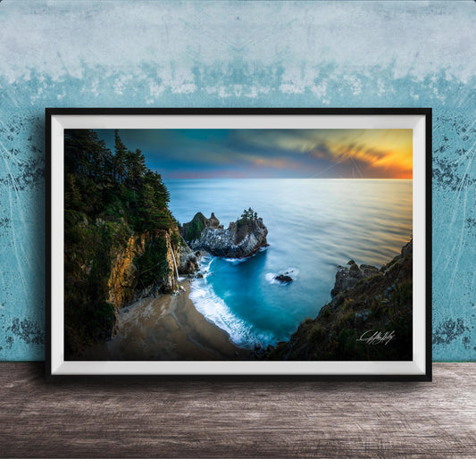 A framed photograph depicting a scenic coastal landscape with a rocky shoreline, a sandy beach, and a body of water with a sunset sky in the background.
