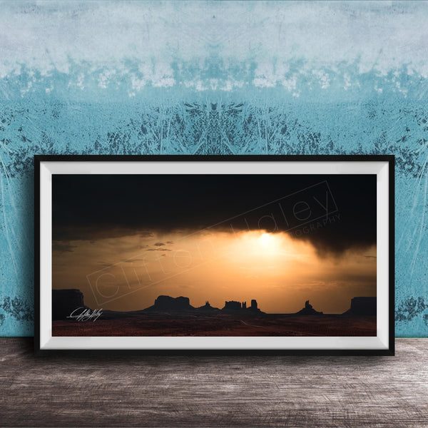 a framed photograph of a desert landscape at sunset, with silhouetted mountains in the background and a dramatic sky filled with clouds and the sun setting behind them.