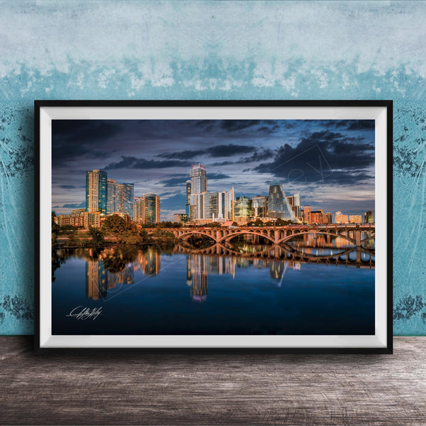A framed photograph of a city skyline at sunset, with the buildings reflected in the water below.