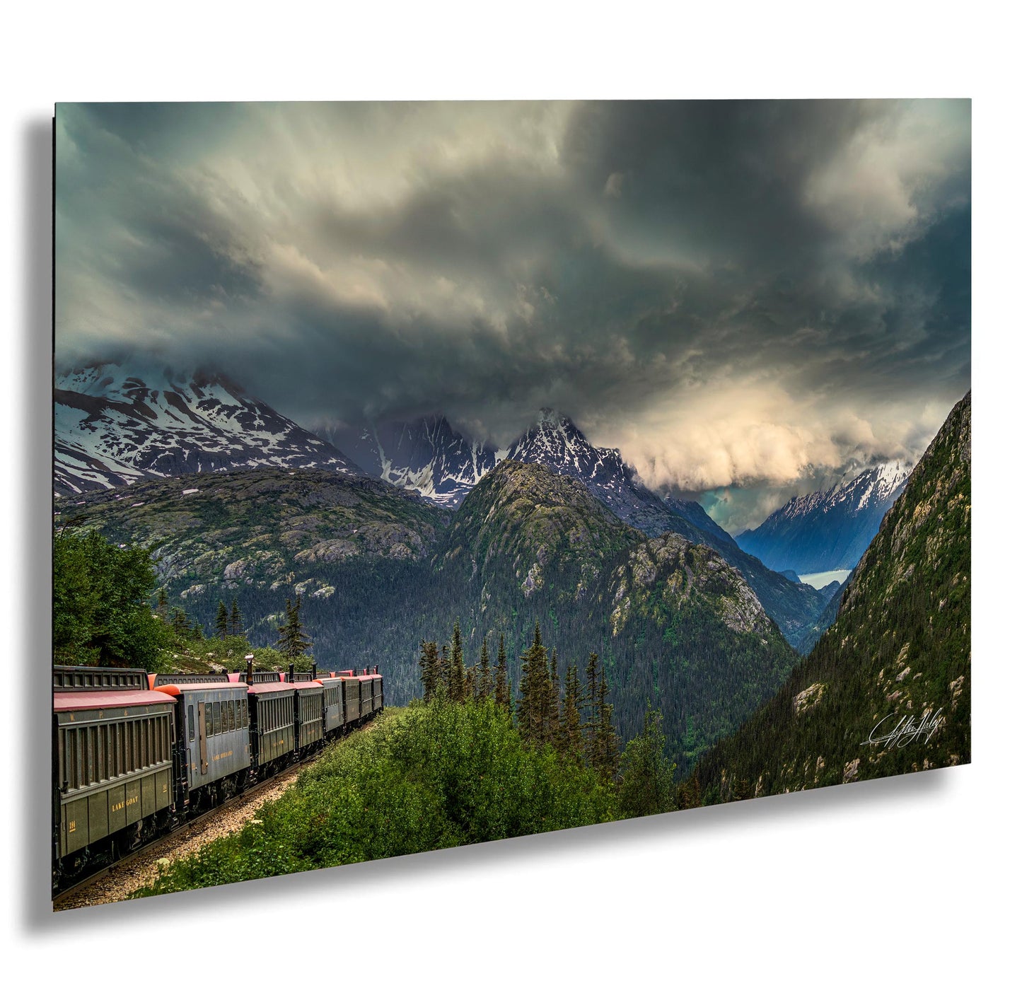 A train travels through a mountainous landscape, with snow-capped peaks and a dramatic cloudy sky in the background.