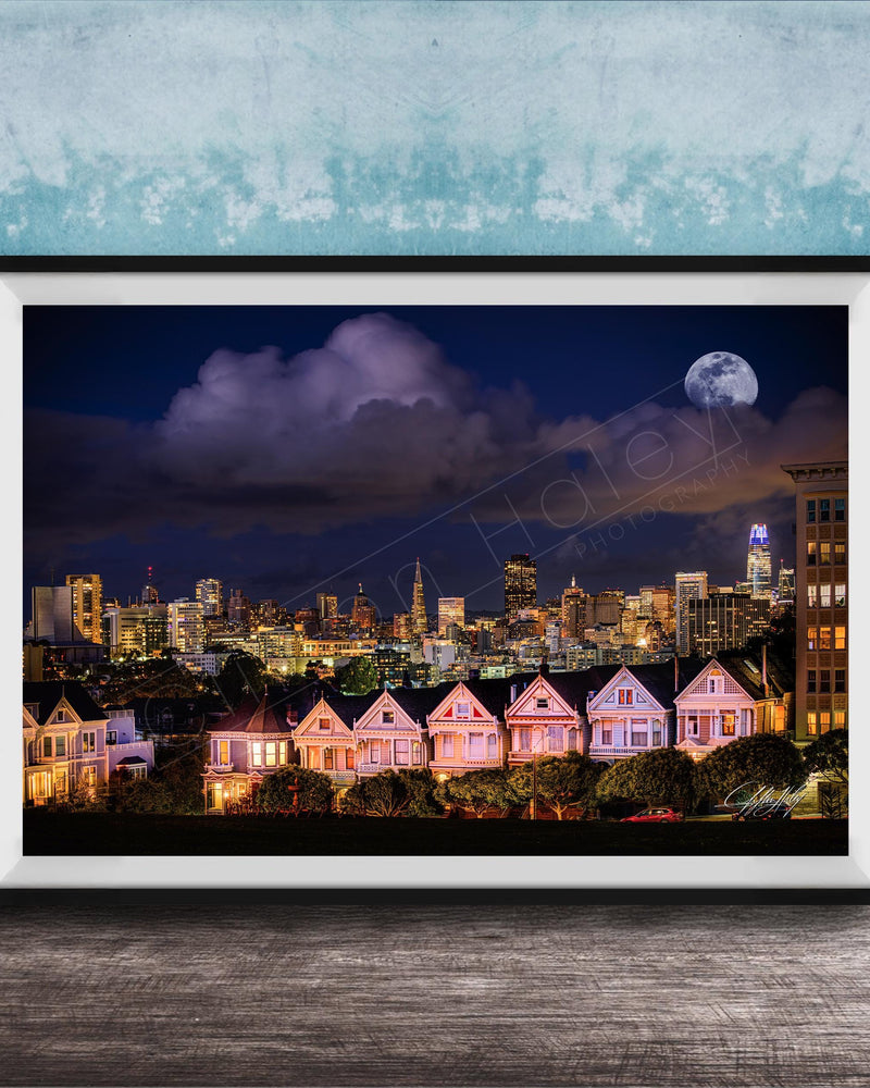 A framed photograph of a city skyline at night, with a full moon in the sky and colorful houses lining the streets.