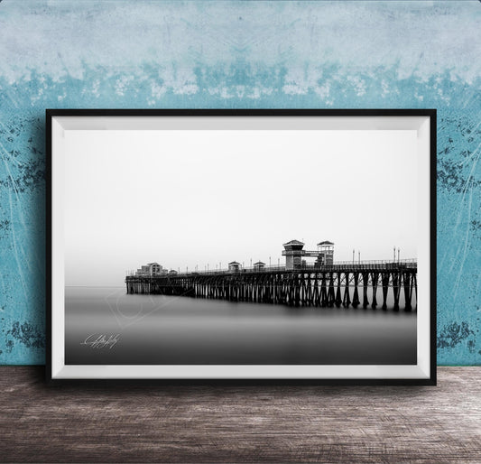 A black and white photograph of a long pier extending into the distance, with a calm body of water below.