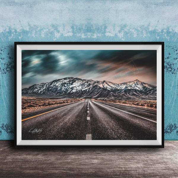 A framed photograph depicting a long, straight road leading towards a snow-capped mountain range under a dramatic, cloudy sky.