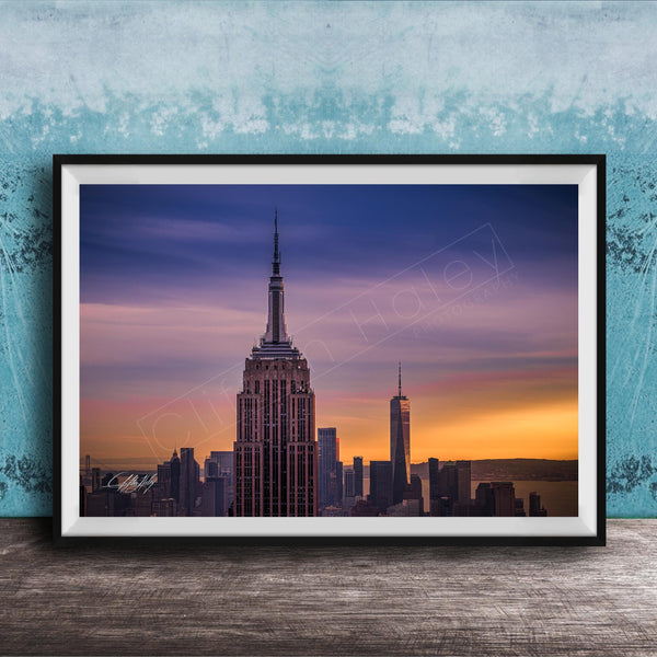 A framed photograph of the New York City skyline at sunset, with the Empire State Building prominently visible in the center of the frame.