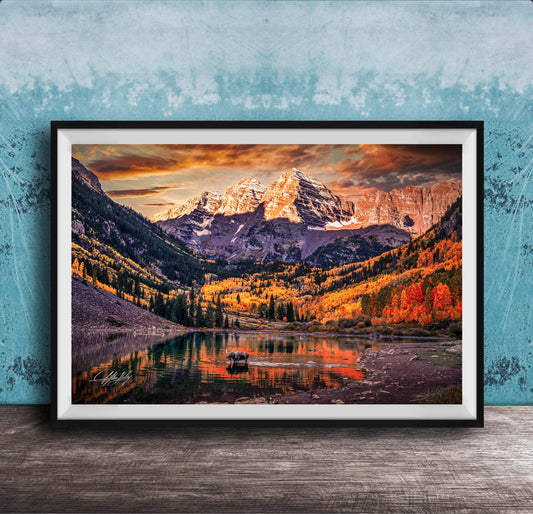 a breathtaking mountain landscape with a lake in the foreground, surrounded by vibrant autumn foliage. The mountains are covered in snow, creating a stunning contrast against the colorful sky. The scene is framed in a black frame and displayed on a wooden surface, adding to the rustic charm of the setting.
