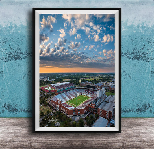 The image is an aerial view of a large sports stadium with a football field, surrounded by buildings and a cloudy sky.