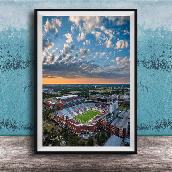 The image is an aerial view of a large sports stadium with a football field, surrounded by buildings and a cloudy sky.