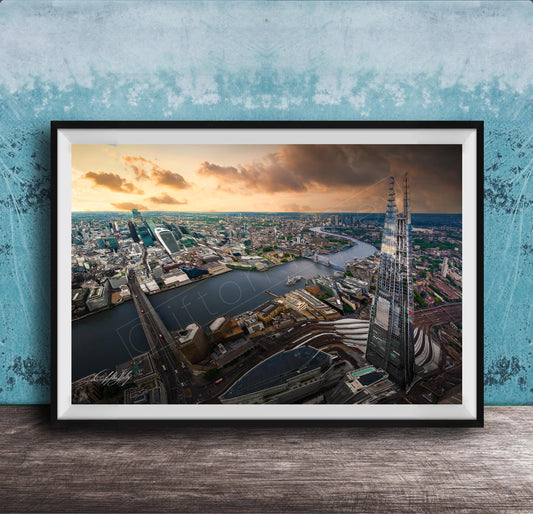 The image is a framed aerial view of a cityscape, showcasing a mix of tall buildings, roads, and a river. The sky is filled with clouds, creating a dramatic backdrop for the city's skyline. The image is framed and displayed on a wooden surface, likely a wall or shelf.