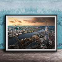 The image is a framed aerial view of a cityscape, showcasing a mix of tall buildings, roads, and a river. The sky is filled with clouds, creating a dramatic backdrop for the city's skyline. The image is framed and displayed on a wooden surface, likely a wall or shelf.