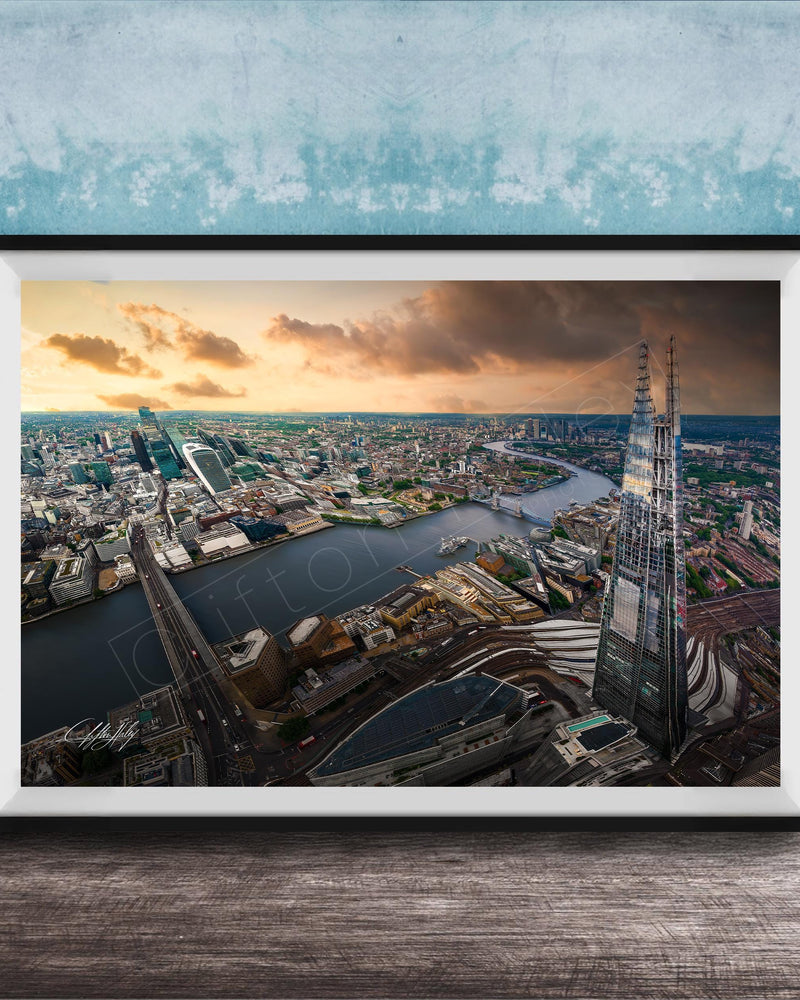 The image is a framed aerial view of a cityscape, showcasing a mix of tall buildings, roads, and a river. The sky is filled with clouds, creating a dramatic backdrop for the city's skyline. The image is framed and displayed on a wooden surface, likely a wall or shelf.
