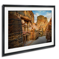 a picturesque canyon with ancient ruins, including a temple carved into the rock face. The canyon walls are adorned with layers of rock, showcasing the geological history of the area. The sky above is a clear blue, providing a beautiful backdrop to the scene. The image is framed and appears to be a photograph, capturing the natural beauty of the landscape.