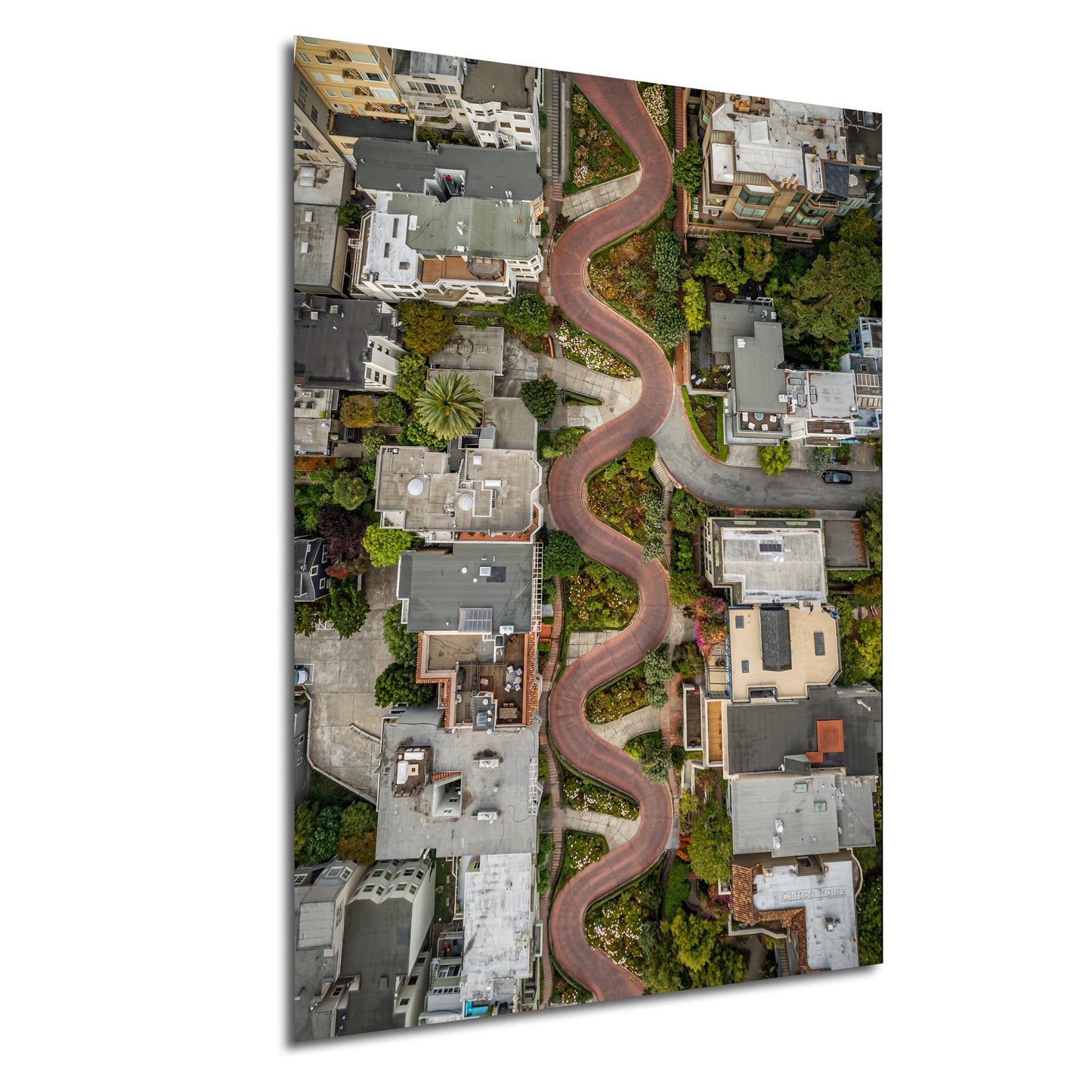The image is an aerial view of a winding road that curves through a densely populated urban area, with buildings and greenery lining the sides of the road.