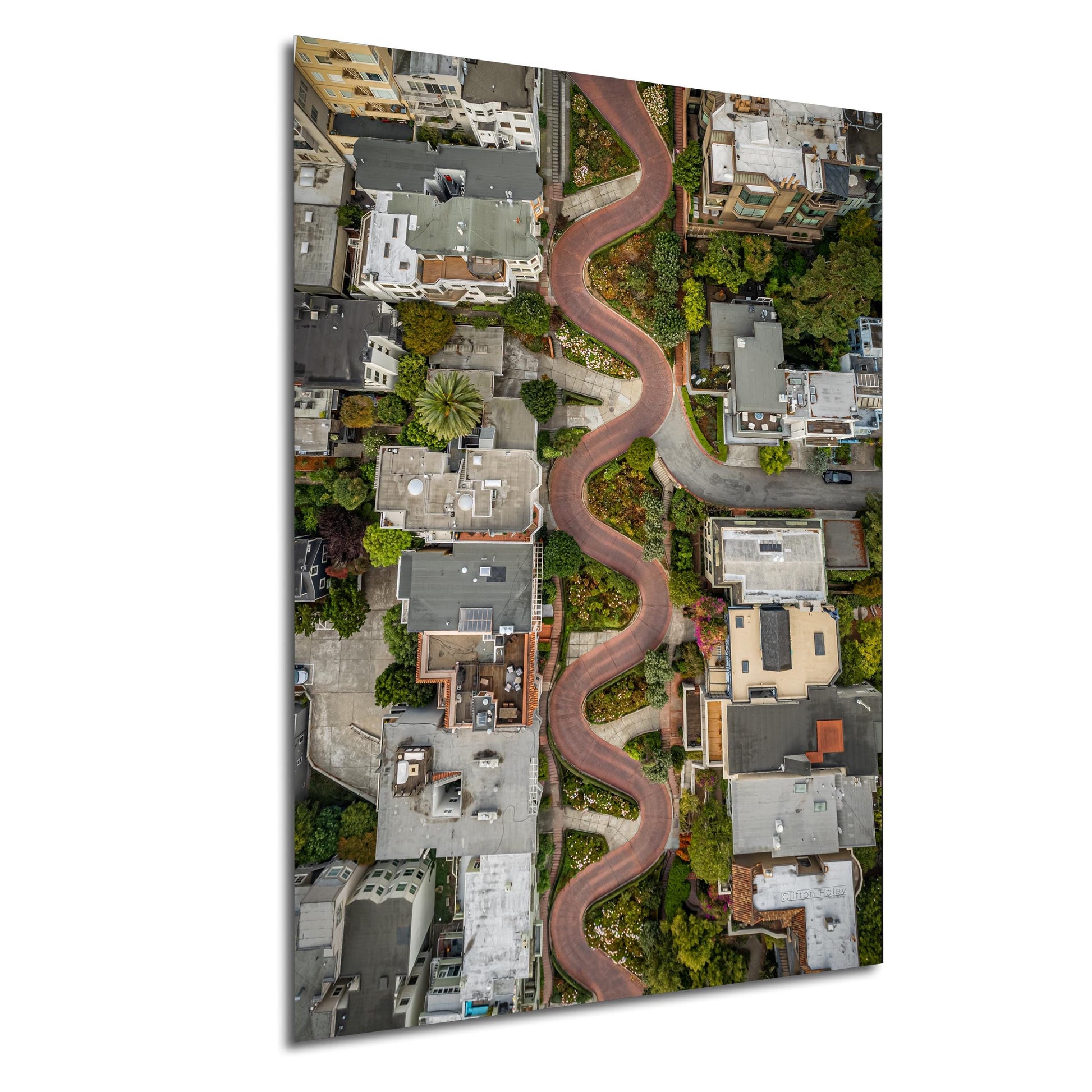 The image is an aerial view of a winding road that curves through a densely populated urban area, with buildings and greenery lining the sides of the road.