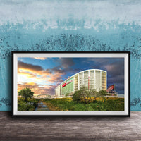 A framed photograph of a large building with a curved roof, surrounded by trees and a river, with a colorful sunset sky in the background.