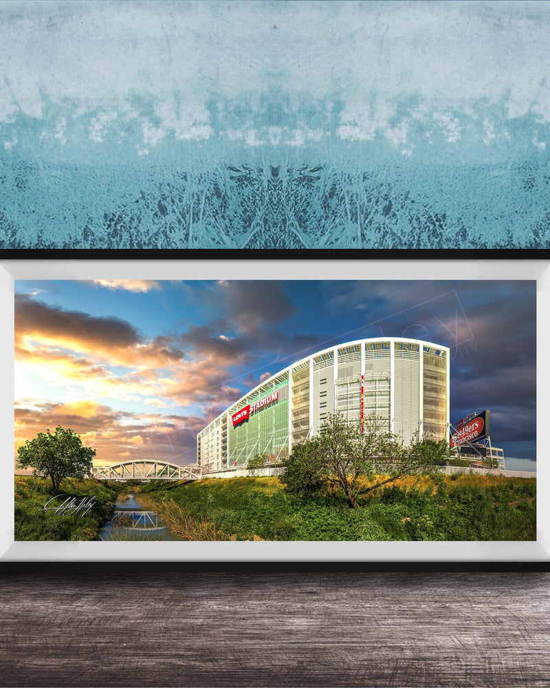A framed photograph of a large building with a curved roof, surrounded by trees and a river, with a colorful sunset sky in the background.