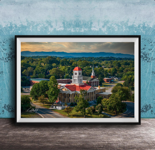 A framed aerial view of a historic building with a red roof, surrounded by lush greenery and mountains in the distance.