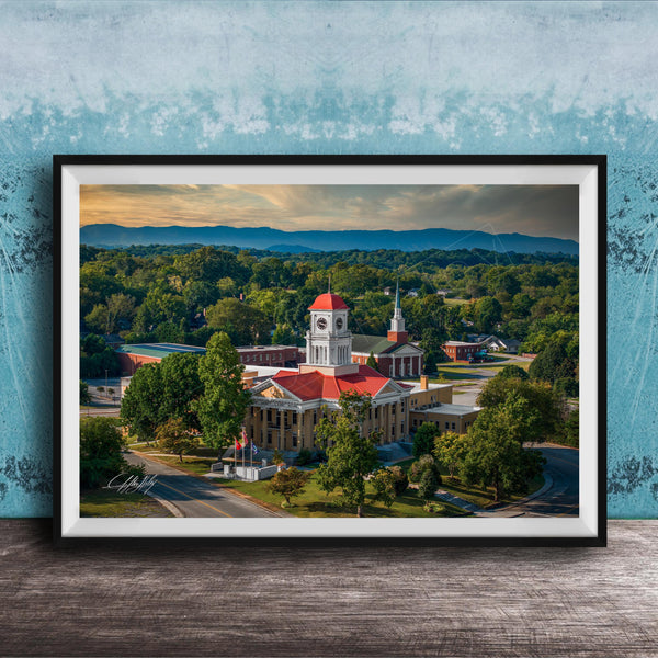 A framed aerial view of a historic building with a red roof, surrounded by lush greenery and mountains in the distance.