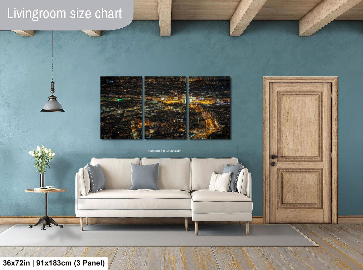 The image is a living room with a large, three-panel wall art depicting a cityscape at night. The wall art is mounted above a white sofa, and there is a wooden door and a small table with a vase of flowers nearby. The living room has a blue wall and a wooden floor.