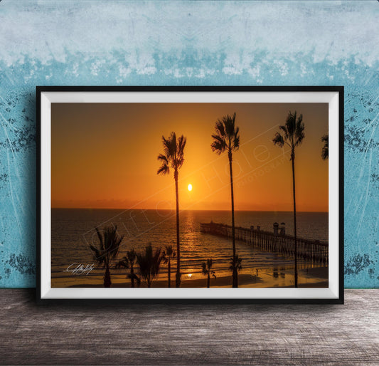 A framed photograph of a serene beach scene at sunset, with palm trees silhouetted against the warm orange sky.