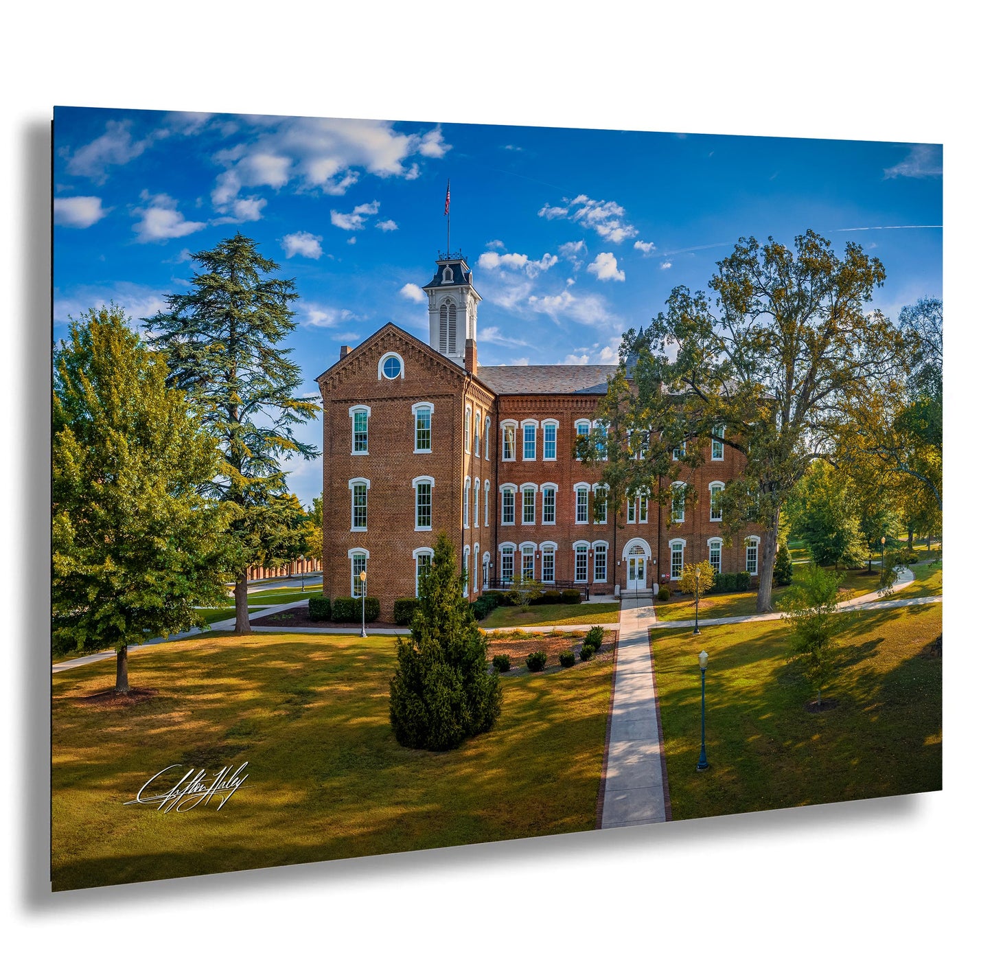 A large brick building with a clock tower, surrounded by trees and a grassy lawn, set against a blue sky with scattered clouds.