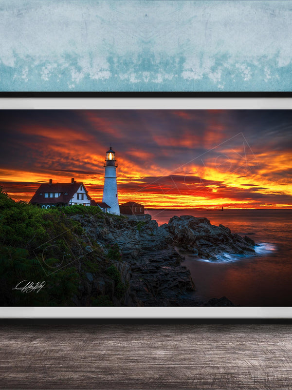 A framed photograph depicting a lighthouse on a rocky cliff at sunset, with the sky filled with vibrant orange and red hues.