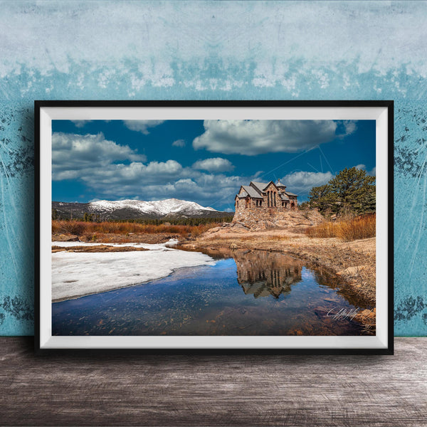 A framed photograph depicting a stone building reflected in a calm body of water, with snow-capped mountains in the background and a cloudy blue sky above.