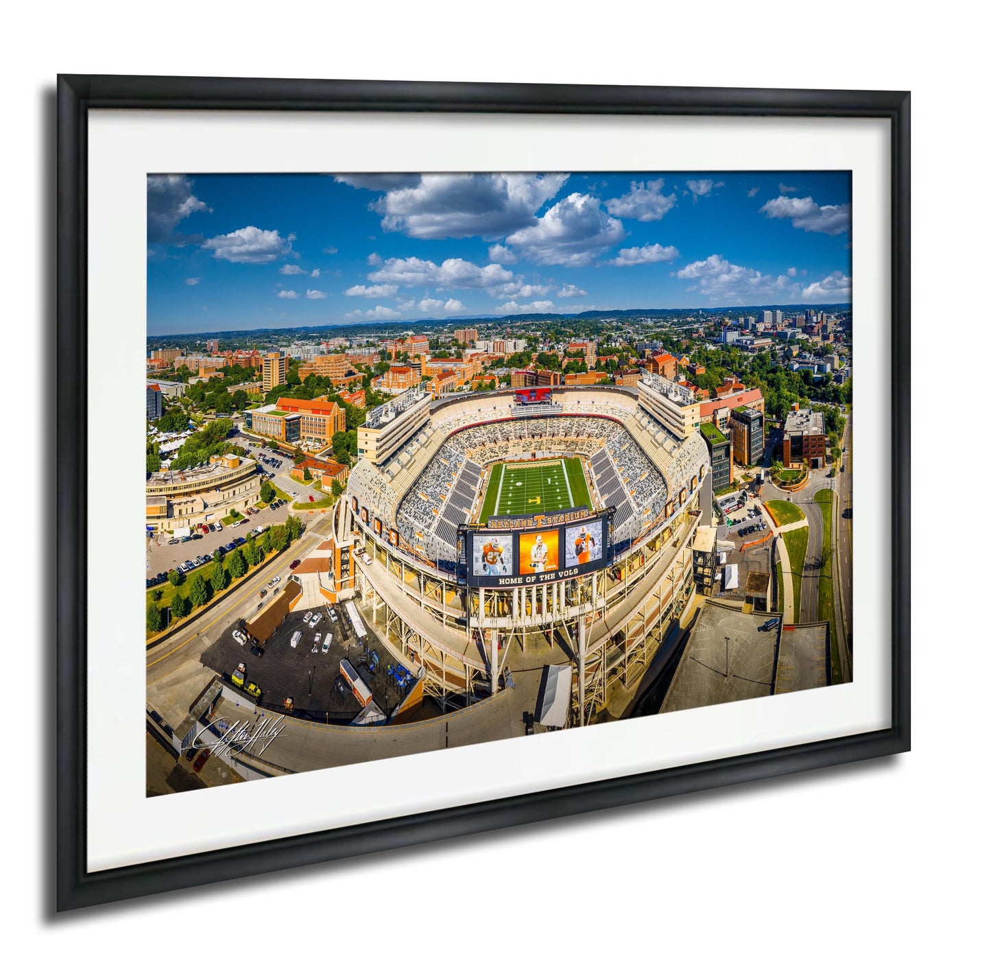 An aerial view of a large sports stadium surrounded by a cityscape, with a clear blue sky and scattered clouds above.