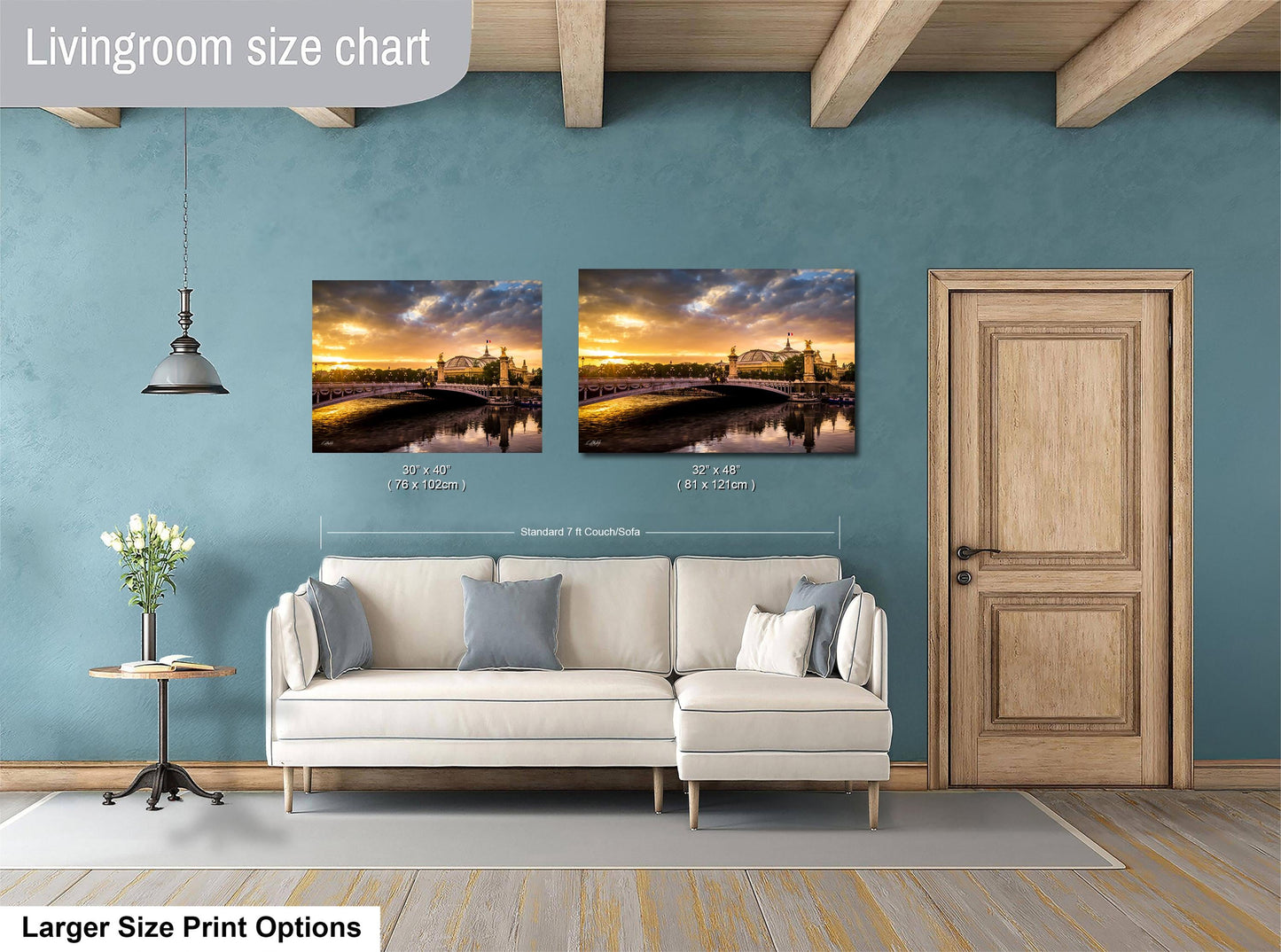 a living room with a white sofa and a wooden door. Above the sofa, there are two framed pictures of a scenic landscape, likely depicting a sunset or a river. The living room has a blue wall and a wooden ceiling, creating a cozy and inviting atmosphere.