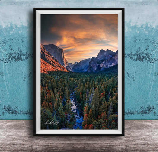 The image is a framed photograph of a mountainous landscape with a river running through it. The mountains are covered in trees, and the sky is filled with clouds, creating a dramatic and colorful scene.