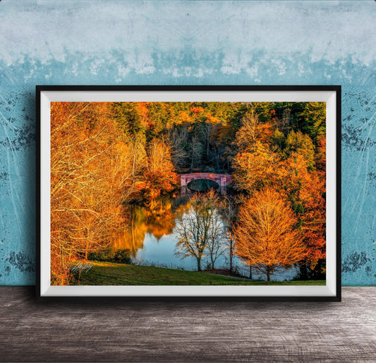 A framed photograph depicting a scenic autumn landscape with a red bridge spanning a tranquil lake surrounded by vibrant orange and yellow foliage.