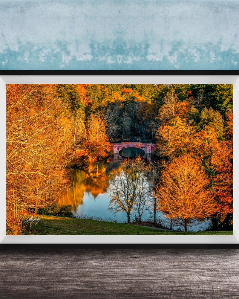 A framed photograph depicting a scenic autumn landscape with a red bridge spanning a tranquil lake surrounded by vibrant orange and yellow foliage.