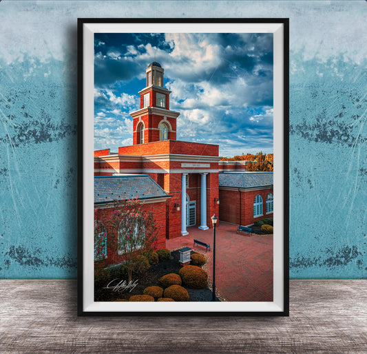 A framed photograph of a red brick building with a tall tower, set against a blue sky with clouds.
