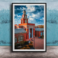 A framed photograph of a red brick building with a tall tower, set against a blue sky with clouds.