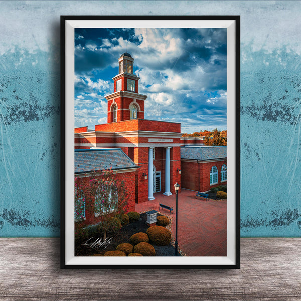 A framed photograph of a red brick building with a tall tower, set against a blue sky with clouds.