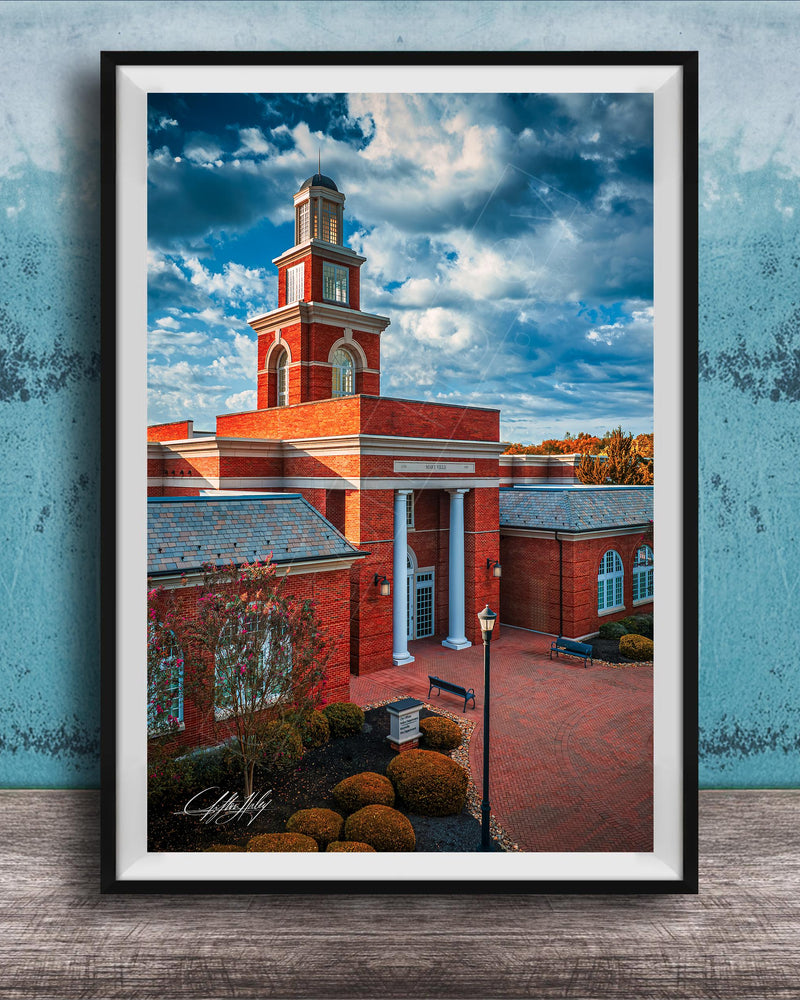 A framed photograph of a red brick building with a tall tower, set against a blue sky with clouds.