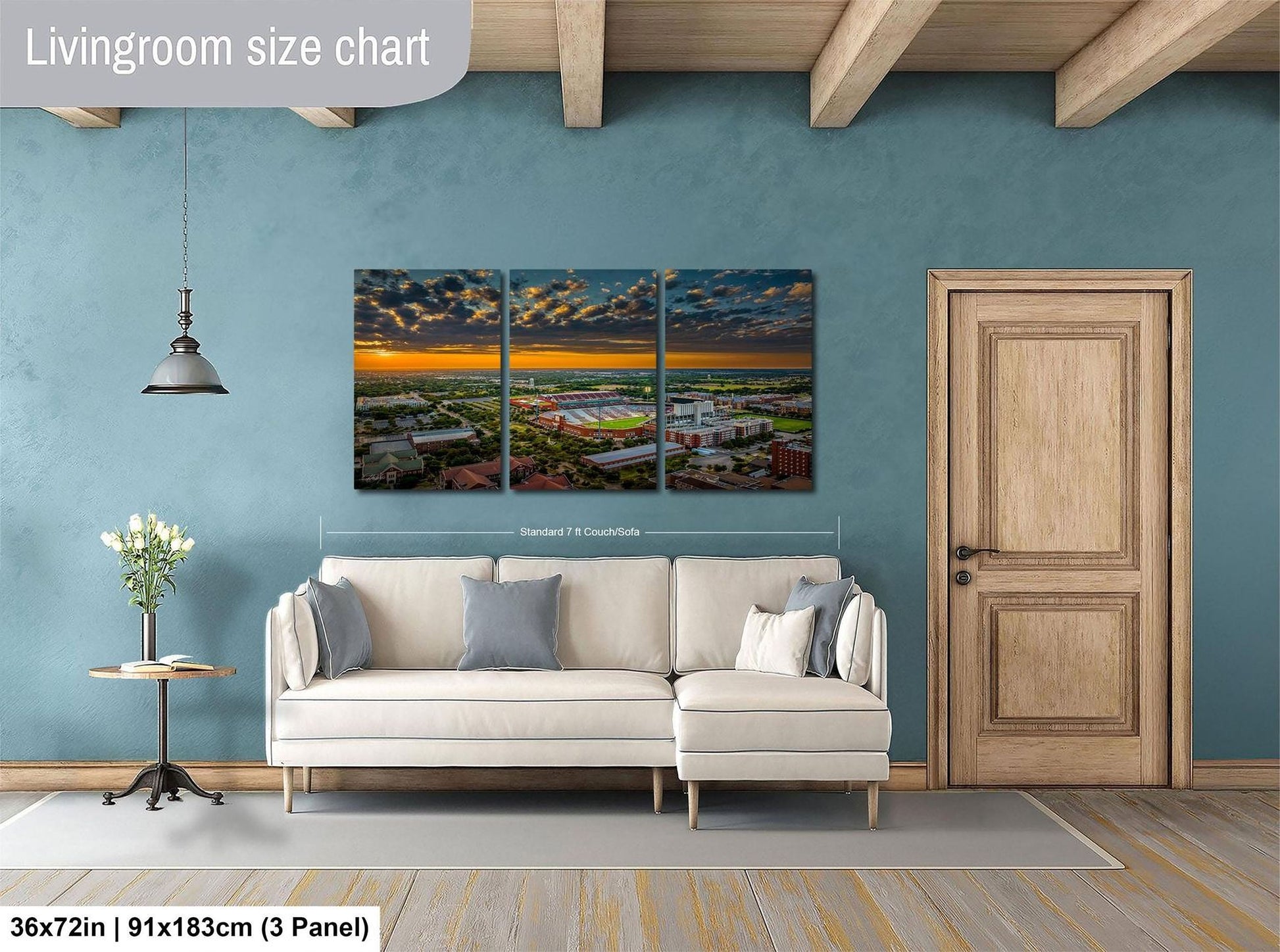 The image is a living room with a large wall art depicting a cityscape at sunset. The wall art is divided into three panels, with the largest panel spanning approximately 36 square inches. The room also includes a white sofa, a wooden door, and a small table with a vase of flowers.
