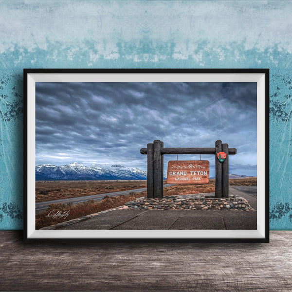 A framed photograph of a sign for Grand Teton National Park, with a cloudy sky and mountains in the background.