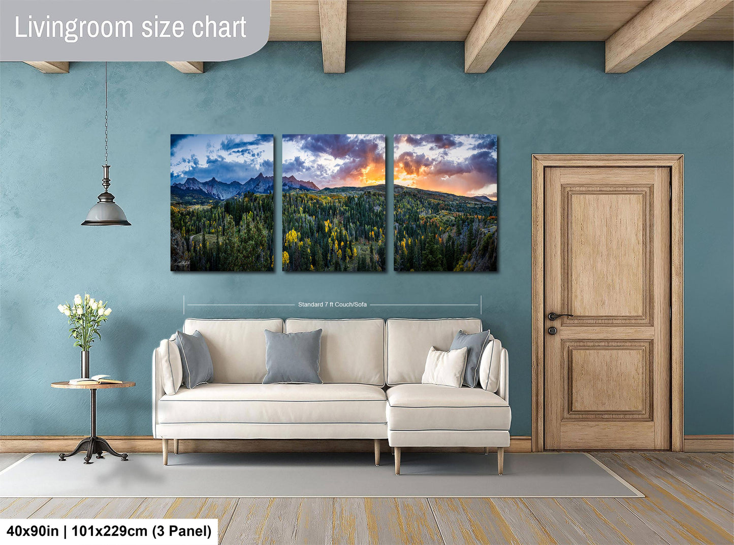 The image is a living room with a large wall art panel depicting a scenic landscape with mountains, trees, and a colorful sky. The panel is divided into three smaller panels, each showing a different part of the landscape. The living room has a white sofa and a wooden door, and there is a potted plant and a vase on a table nearby. The wall art panel is approximately 40 inches wide and 101.29 cm tall, and it is mounted on the wall.
