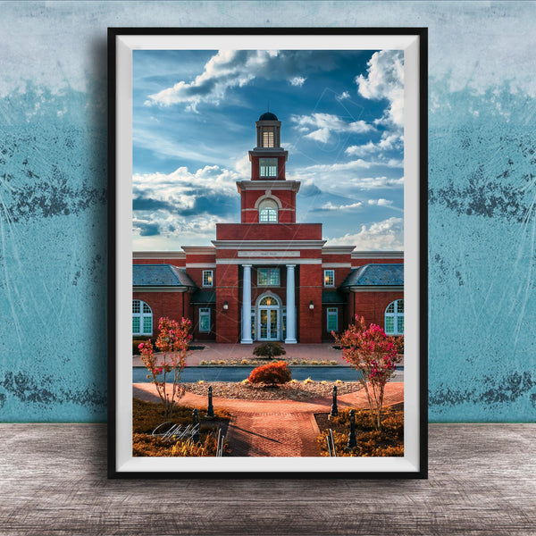 A framed photograph of a red brick building with a prominent clock tower, set against a blue sky with clouds.