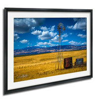 A windmill stands tall in a vast, golden field under a blue sky dotted with fluffy white clouds.