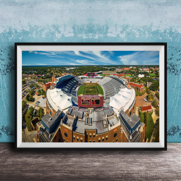 The image is an aerial view of a large sports stadium with a football field, surrounded by buildings and a blue sky.