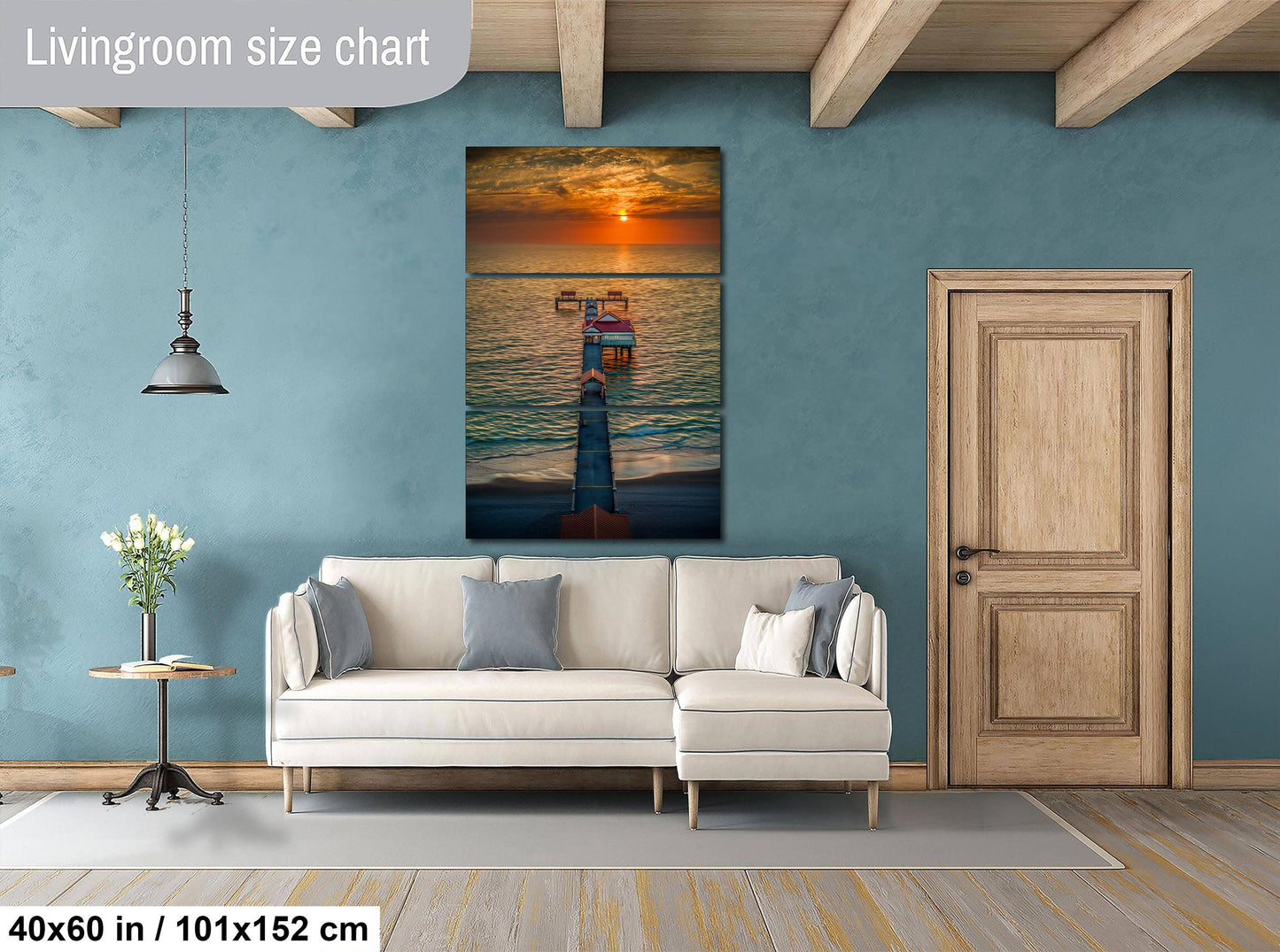 a living room with a large wall painting of a sunset over the ocean, a white sofa, a wooden door, and a small table with a vase of flowers.