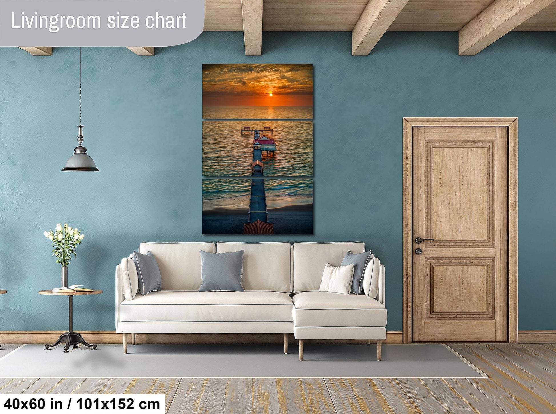 a living room with a large wall painting of a sunset over the ocean, a white sofa, a wooden door, and a small table with a vase of flowers.