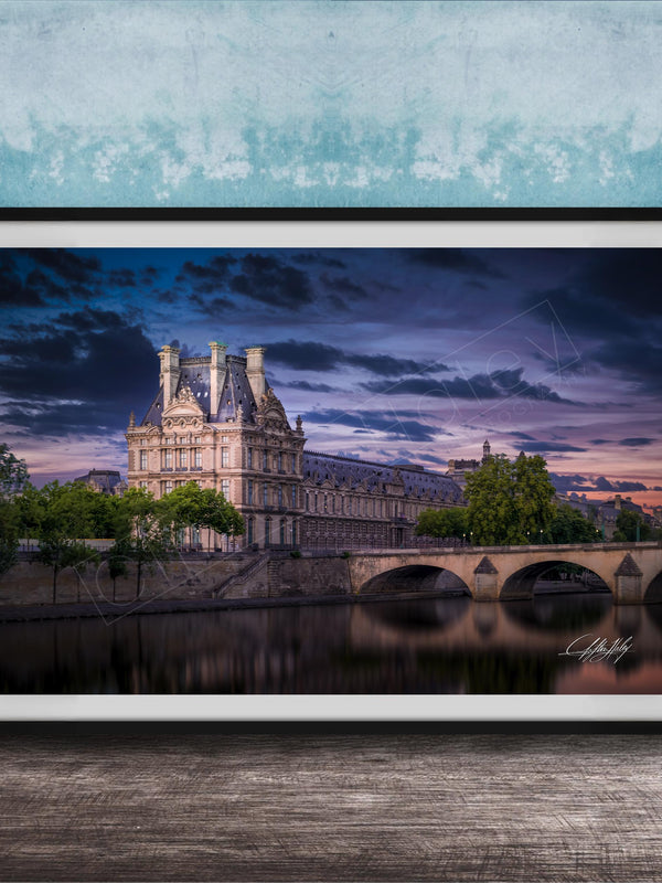 a large, ornate building with a tower and multiple spires, situated on the banks of a river. The sky is filled with clouds, creating a dramatic backdrop for the scene. The building is surrounded by trees, and a bridge can be seen in the background. The image is framed and displayed on a wooden surface, with a blue wall in the background.