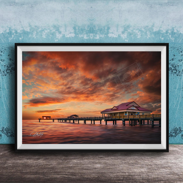 A framed photograph depicting a serene sunset over a wooden pier, with a small building on the pier and a calm body of water in the foreground.