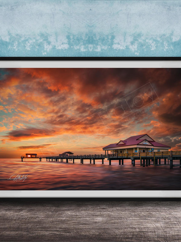 A framed photograph depicting a serene sunset over a wooden pier, with a small building on the pier and a calm body of water in the foreground.
