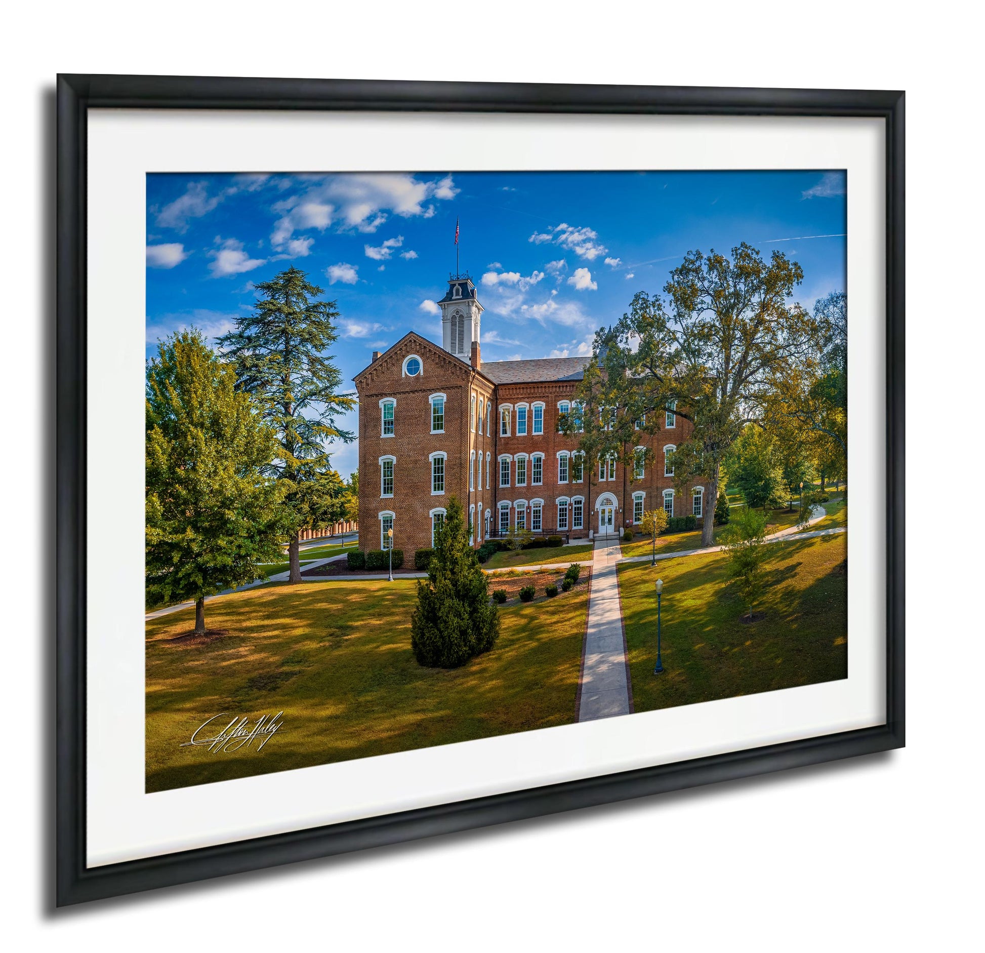 A brick building with a tower and a flag on top, surrounded by trees and a grassy area, with a clear blue sky in the background.