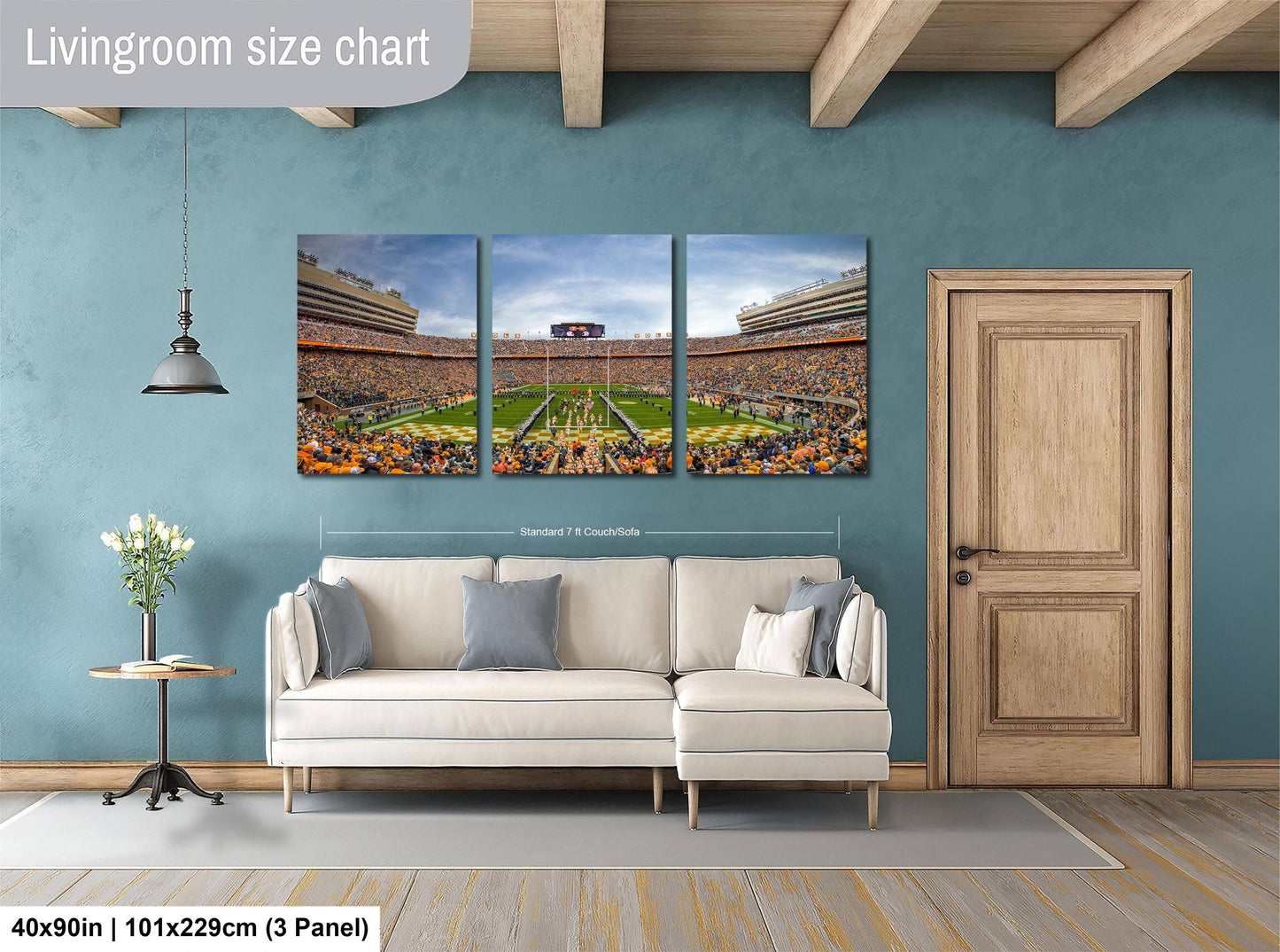 The image is a living room with a large wall-mounted painting depicting a football stadium. The painting is divided into three panels, showing different aspects of the stadium. The room has a white sofa, a wooden door, and a small table with a vase of flowers. The painting is approximately 40 inches wide and 101.29 cm tall, with a 3-panel layout.