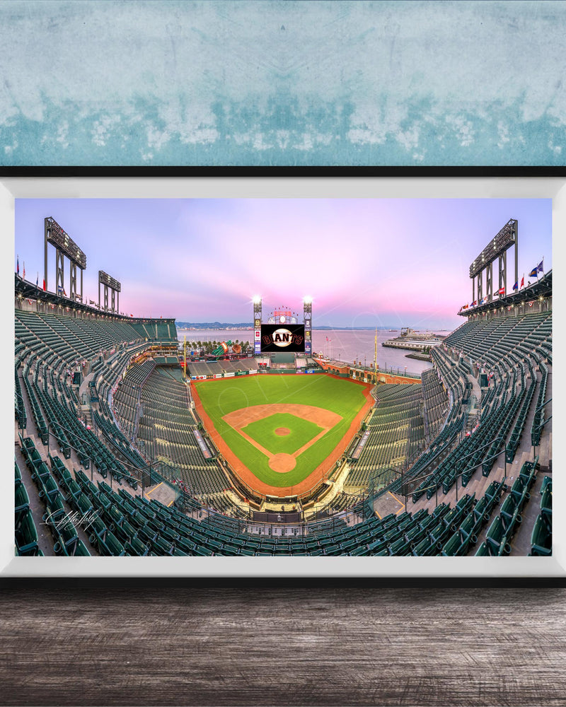 A framed photograph of a baseball stadium at dusk, with the field illuminated and the sky displaying a colorful sunset.