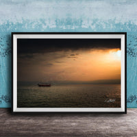 A framed photograph of a boat sailing on a calm body of water at sunset, with a blue sky and clouds in the background.